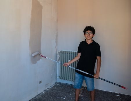 Renovate the walls of the house by painting them: the Caucasian boy with glasses passes the gray impregnated roller on the wall. A little color drips down the wall.