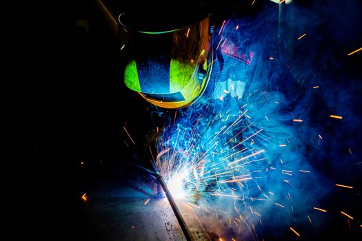 A process of manual operational semi-automatic electric welding with sparks, professional industrial worker in black-yellow protective mask at work.. High contrast dark close-up scene. MIG - metal inert gas - type of welding.