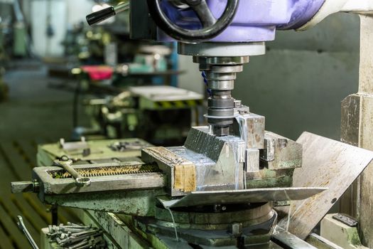 A process of old-style manual milling in weathered vise with liquid coolant., Close-up with selective focus and background bliur, Vertical spindle orientation. Manufacturing theme.