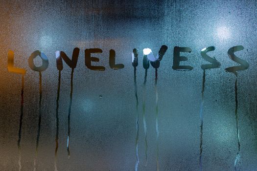 The word loneliness written on night sweaty window glass with blurred blue light in the background and smudges.