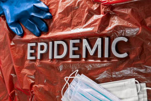 the word epidemic laid by metal letters in crumpled red plastic background with face masks and blue protective glove.