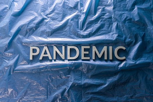 the word pandemic laid with silver letters on crumpled blue plastic film background in flat lay composition at center with dramatic cold light