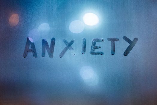 the word anxiety written by finger on night wet glass with blurred classic blue lights in background.