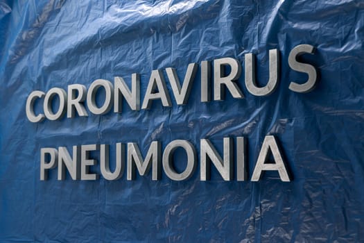 the word coronavirus pneumonia laid with silver letters on crumpled blue plastic film in side perspective composition with dramatic light
