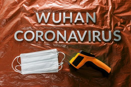 words wuhan coronavirus laid with metal letters on orange crumpled plastic film backdrop with face masks and infrared thermometer - flat lay with dramatic light