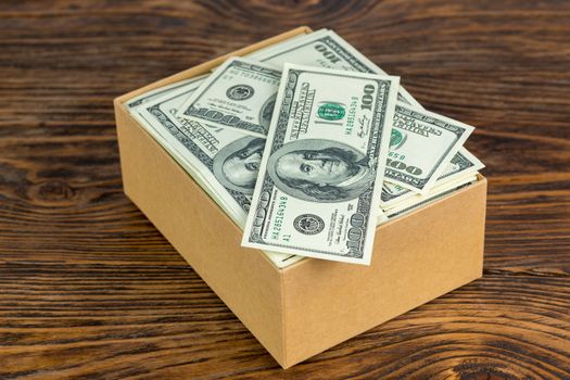 small cardboard box full of hundred dollar banknotes close-up with selective focus on natural wood background.