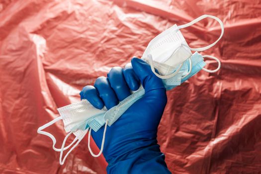 A hand in blue medical glove holding a bunch of face masks on blurry red plastic film background. Symbol of coronavirus pandemic resistance and help.