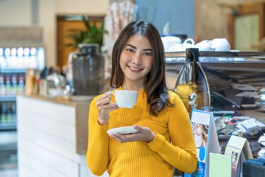 Asian young female holding a cup of coffee in modern coffee shop or coworking space beside coffee espresso machine, Lifestyle and leisure with hobby and entrepreneur concept