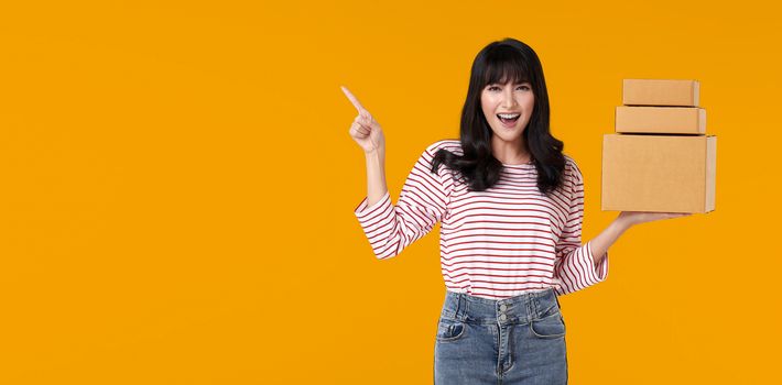 Happy casual asian woman carrying a delivery box that has been packaged for shipping. ready to point  finger in the blank space isolated on bright orange background.