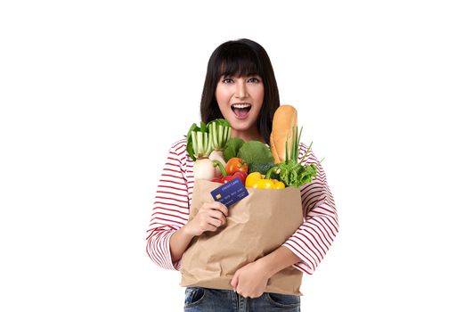 Happy Asian woman with credit card holding paper bag full of fresh vegetable groceries isolated on white background.