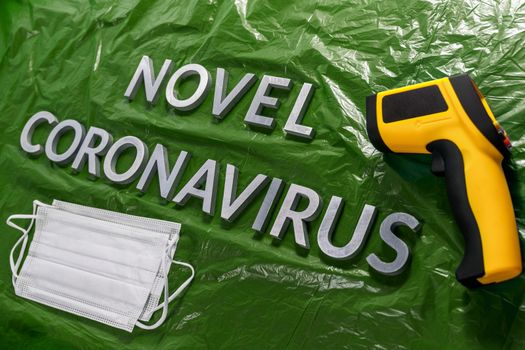 words novel coronavirus laid with metal letters on green crumpled plastic film backdrop with face masks and infrared thermometer - diagonal perspective with dramatic light