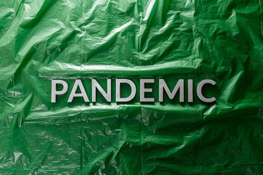 the word pandemic laid with silver letters on green crumpled plastic film background in flat lay composition at center with dramatic cold light