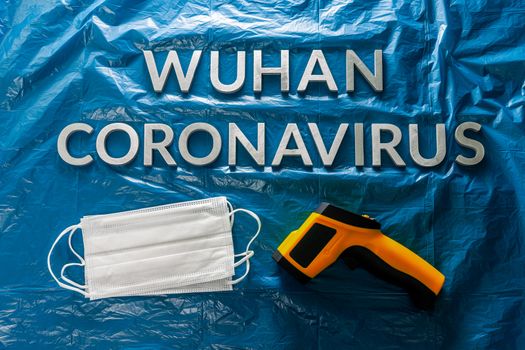 words wuhan coronavirus laid with metal letters on crumpled blue plastic film backdrop with face masks and infrared thermometer - flat lay with dramatic light