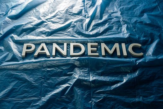 The word pandemic laid with silver letters on crumpled blue plastic film background Slanted centered with dramatic cold light.