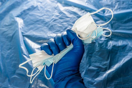 A hand in blue medical glove holding a bunch of face masks on blurry plastic film background. Symbol of coronavirus pandemic resistance and help.