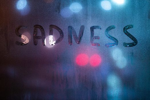 the word sadness written by finger on night wet glass with blurred blue and red lights in background.