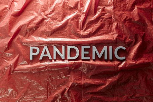 the word pandemic laid with silver letters on red crumpled plastic film background in flat lay composition at center with dramatic cold light