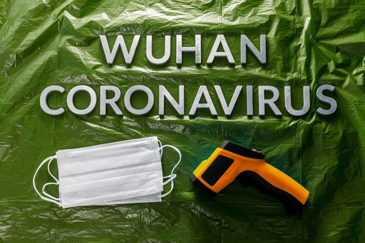 words wuhan coronavirus laid with metal letters on green crumpled plastic film backdrop with face masks and infrared thermometer - flat lay with dramatic light