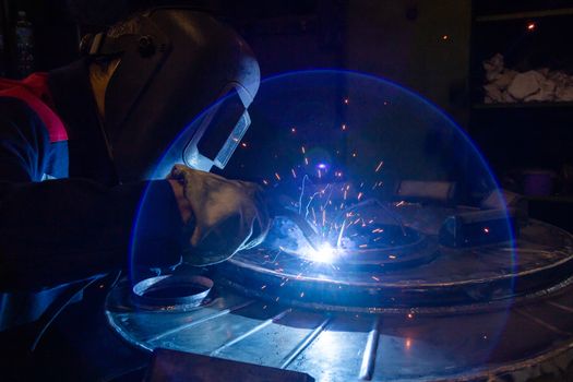 A lens flare while manual semi-automatic electric welding with sparks, professional industrial worker in black protective mask at work. High contrast dark close-up scene. MIG - metal inert gas - type of welding.