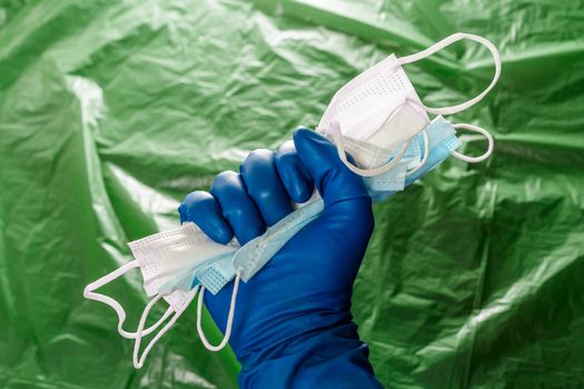 A hand in blue medical glove holding a bunch of face masks on blurry green plastic film background. Symbol of coronavirus pandemic resistance and help.