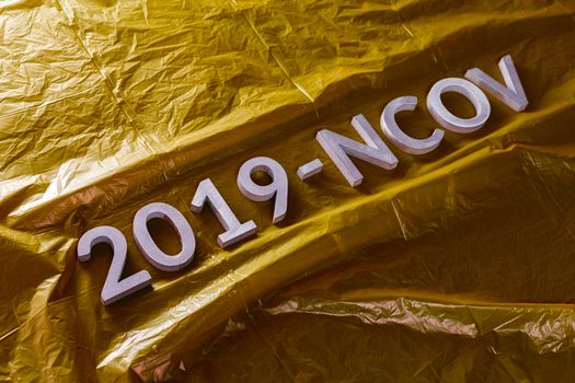 The word 2019-NCOV laid with metal letters on yellow crumpled plastic film background in slanted diagonal composition with dramatic light. Abstract chinese coronavirus concept,