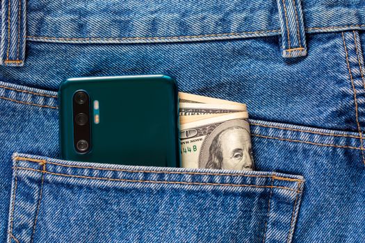 US dollar banknotes and back side of a green smartphone in the left rear pocket of blue jeans. Concept of saving money or pocket expenses.