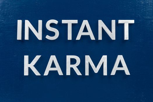 the phrase instant karma laid with silver metal letters on classic blue painted board background.