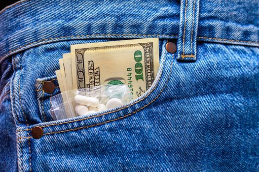 White drug pills and US dollar banknotes in the front left pocket of blue jeans. Concept of medical expenses or illegal drug dealing