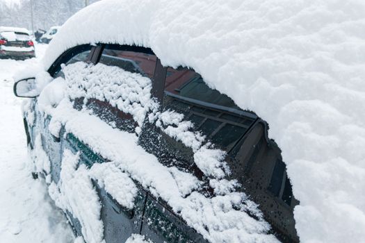 Parked car covered by thick snow layer after winter blizzard at cloudy day light - close-up with selective focus and blur.