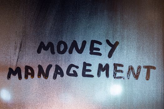 the words money management handwritten on night wet window glass with selective focus and blur in background.