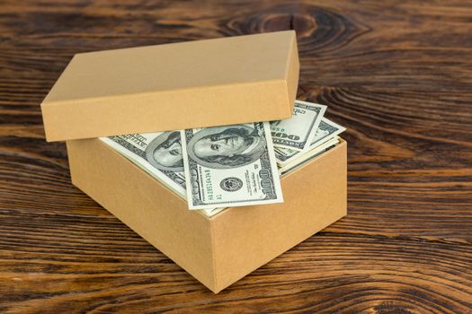 small cardboard box full of hundred dollar banknotes close-up with selective focus on natural wood background.