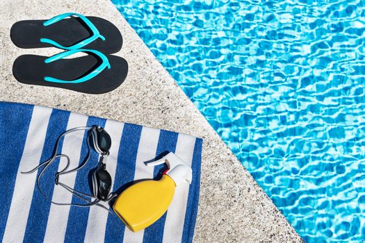 Flip flops, sunscreen spray and swimming goggles on a blue and white striped towel at the edge of a pool.. Copy space. Top view.