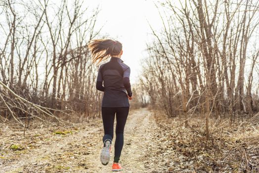 female athlete runner running in autumn nature outdoors on forest trail path in beautiful sunny sunlight fall cold weather. Unrecognizable woman in activewear tights jogging away view from behind.