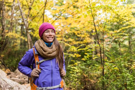 Happy asian woman hiking in autumn forest nature walking on trail path. Hiker girl with backpack, hat, scarf and jacket on fall adventure travel outdoors good weather during hike in cold weather.