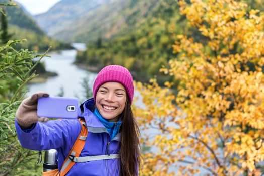 Happy asian hiker woman taking smartphone selfie at scenic viewpoint in nature fall mountain landscape outdoors. Girl hiking in Autumn forest park travel lifestyle. Girl holding phone.