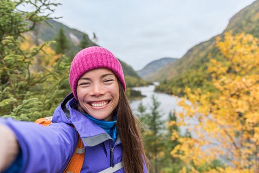 Selfie asian girl hiking in autumn nature mountains. Happy hiker woman taking smartphone picture holding phone at scenic viewpoint in fall mountain landscape outdoors. Forest park travel lifestyle.