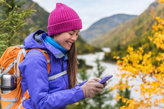 Happy asian girl in cold weather gear hiking in nature using smartphone with touch screen gloves during hike in autumn travel adventure in mountain forest outdoors. Beautiful landscape background.