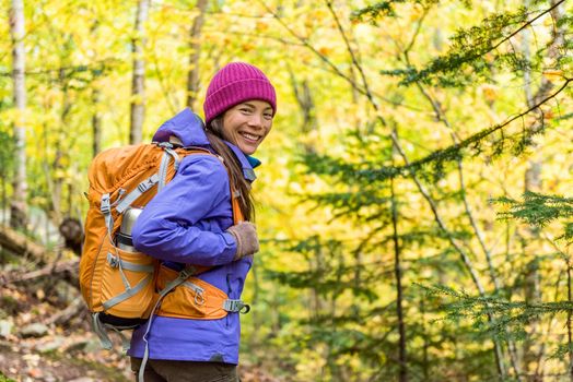 Happy backpacker girl hiking in autumn forest. Young asian hiker woman in outdoor gear for cold weather with backpack looking at camera enjoying walking in nature outdoors in fall season.