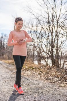 Active woman runner living a modern lifestyle getting ready for trail run on cold autumn forest path in morning nature outdoors. Running healthy girl happy of fitness routine looking at smartwatch.