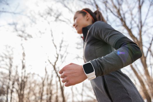 Runner wearing smartwatch on wrist closeup. Young fitness woman running working out cardio in autumn or winter nature outdoors on forest trail in sunlight fall wearing cold weather jacket.