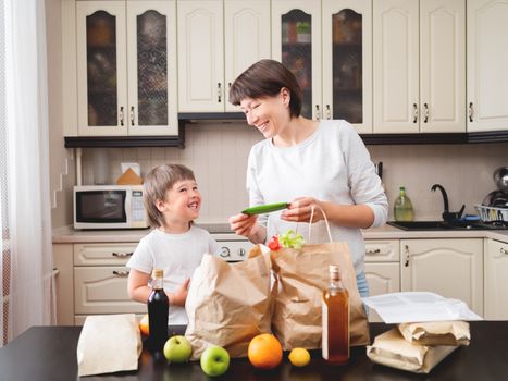 Woman and toddler boys sorts out purchases in the kitchen. Grocery delivery in paper bags. Subscription service from grocery store. Mother and son at kitchen.
