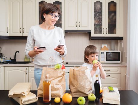 Woman and toddler boys sorts out purchases in the kitchen. Kid bites an apple. Grocery delivery in paper bags. Subscription service from grocery store. Mother and son at kitchen.