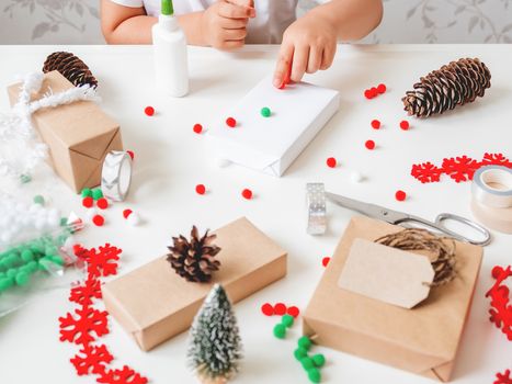 Kid wraps handmade Christmas presents in craft paper with colorful pompons and snowflake ribbons as decorations. Child prepares gifts for New Year celebration. Peaceful leisure activity before winter holidays.