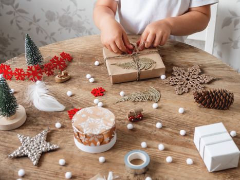 Child wrapped Christmas presents in craft paper with white fluffy pompons and fringe as decoration. Wooden table with hand made New Year gifts.