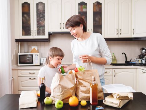 Woman and toddler sorts out purchases in the kitchen. Kid bites muesli bar. Grocery delivery in paper bags. Subscription service from grocery store. Mother and son at kitchen.