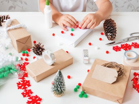 Kid wraps handmade Christmas presents in craft paper with colorful pompons and snowflake ribbons as decorations. Child prepares gifts for New Year celebration. Peaceful leisure activity before winter holidays.