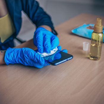 Woman's hand in blue gloves sanitizing cleaning smartphone mobile phone on wood table surface with wet wipes and alcohol