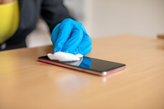 Woman's hand in blue gloves sanitizing cleaning smartphone mobile phone on wood table surface with wet wipes