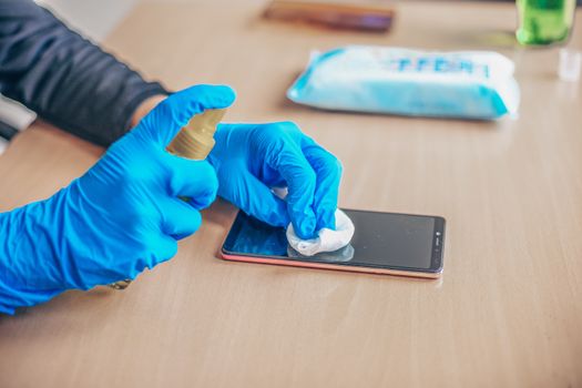 Woman's hand in blue gloves sanitizing cleaning smartphone mobile phone on wood table surface with wet wipes