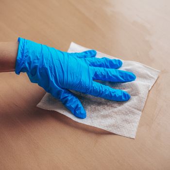 Woman's hand in blue gloves sanitizing cleaning home office wood table surface with wet wipes